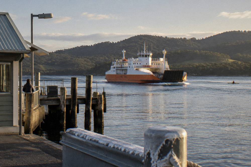 A vehicle ferry mid-crossing on calm grey-green water with forested hills behind