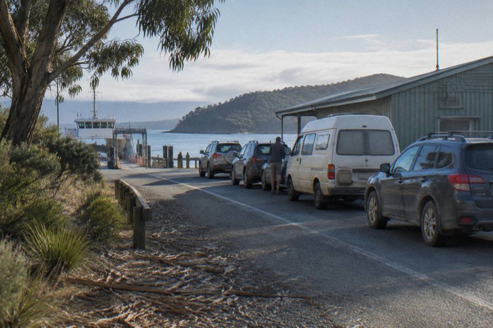 Cars queued in a lane beside a small ferry terminal building waiting for the next sailing