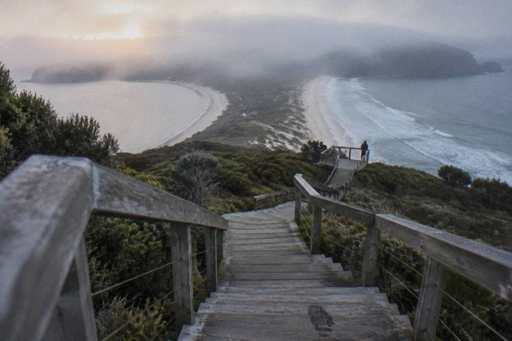 Timber stairs climbing to a lookout platform above a narrow sandy isthmus joining two headlands