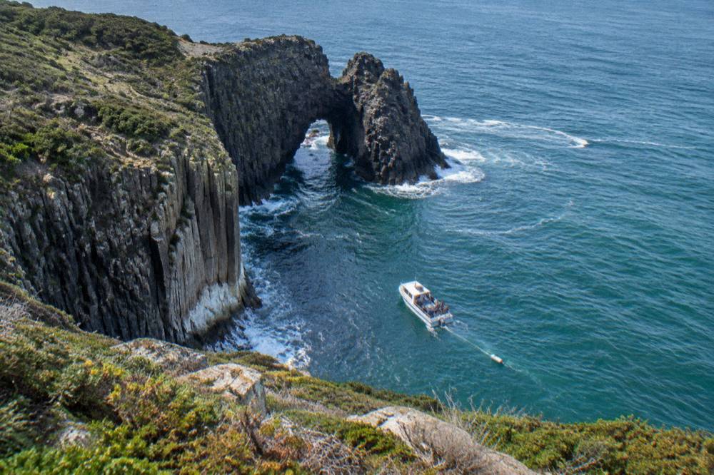 A small open cruise boat riding swell beneath tall sea cliffs and a natural rock arch