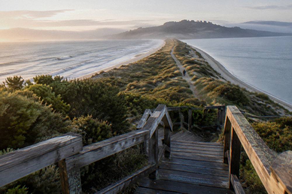 Viewing platform and wooden steps over grassed dunes leading to a narrow sand isthmus between two island halves