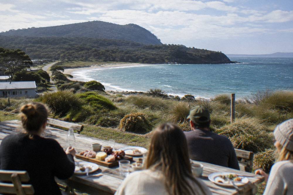 A tour group seated at a long wooden table outdoors, eating lunch with a view of the bay behind them
