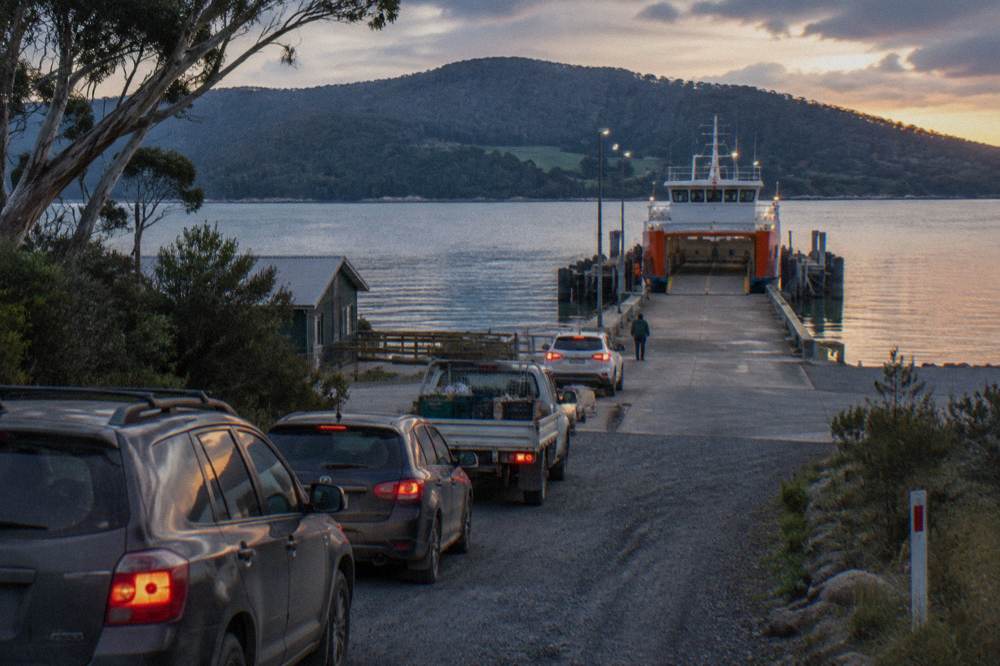 A small car ferry with a white vehicle queue waiting on the Kettering ramp, forested hills behind