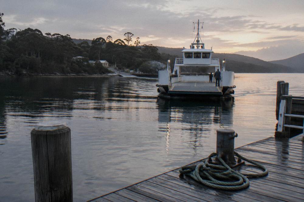 A white vehicle ferry approaching a small timber jetty across calm water