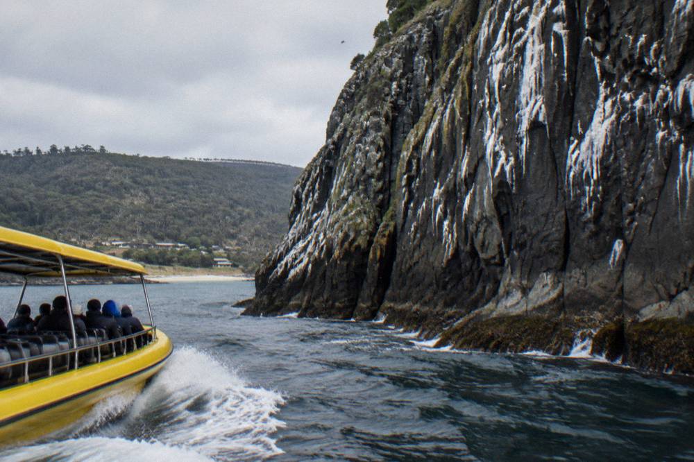 Open cruise boat running close beneath tall sea cliffs on a wild coastline