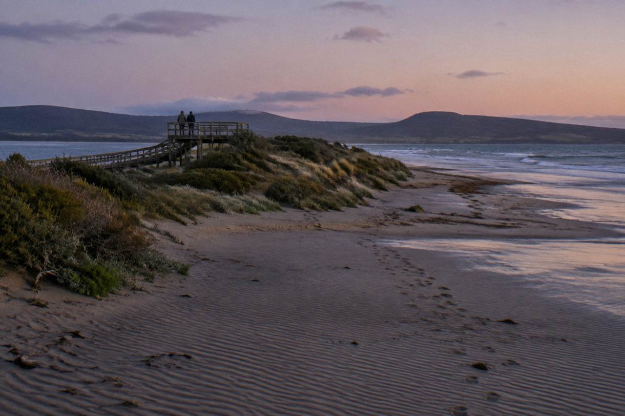 Wooden boardwalk and viewing platform above a narrow sand spit at dusk, with low coastal hills on the horizon