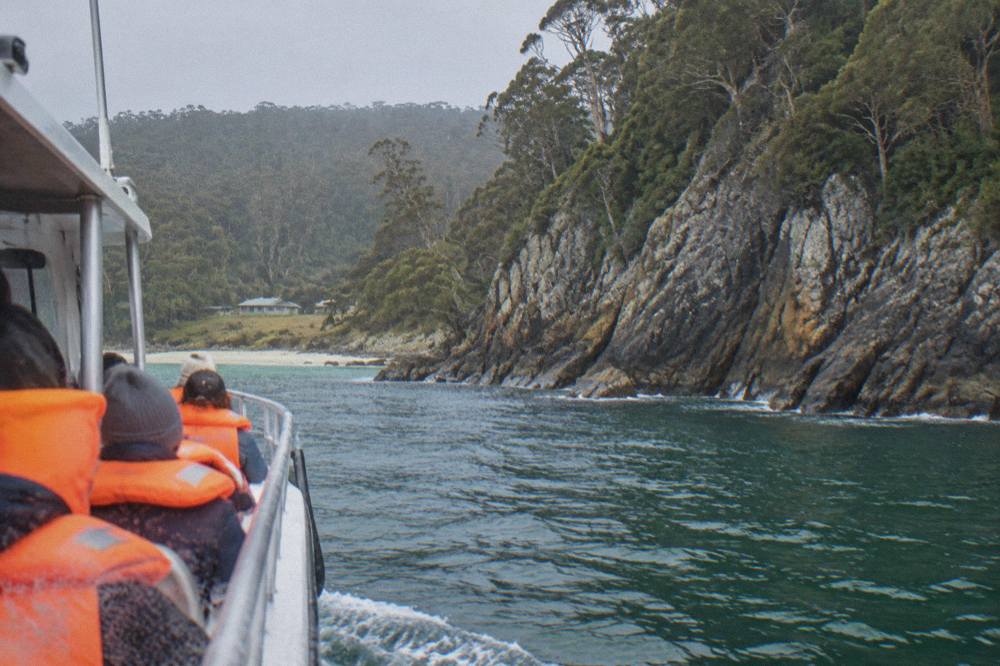 An open cruise boat travelling close to a steep rocky shoreline with deep green water