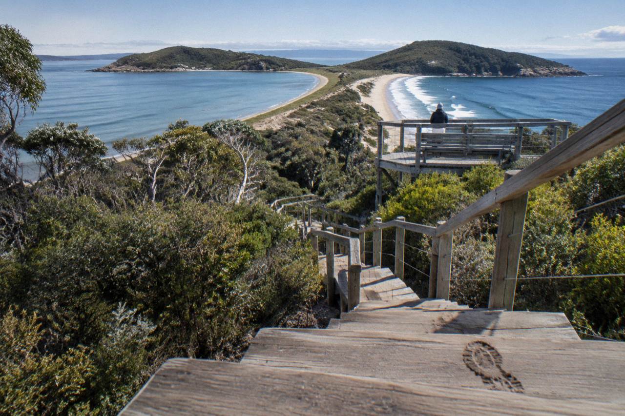 A viewing platform and wooden steps above a narrow sandy isthmus joining two green island headlands under a wide sky