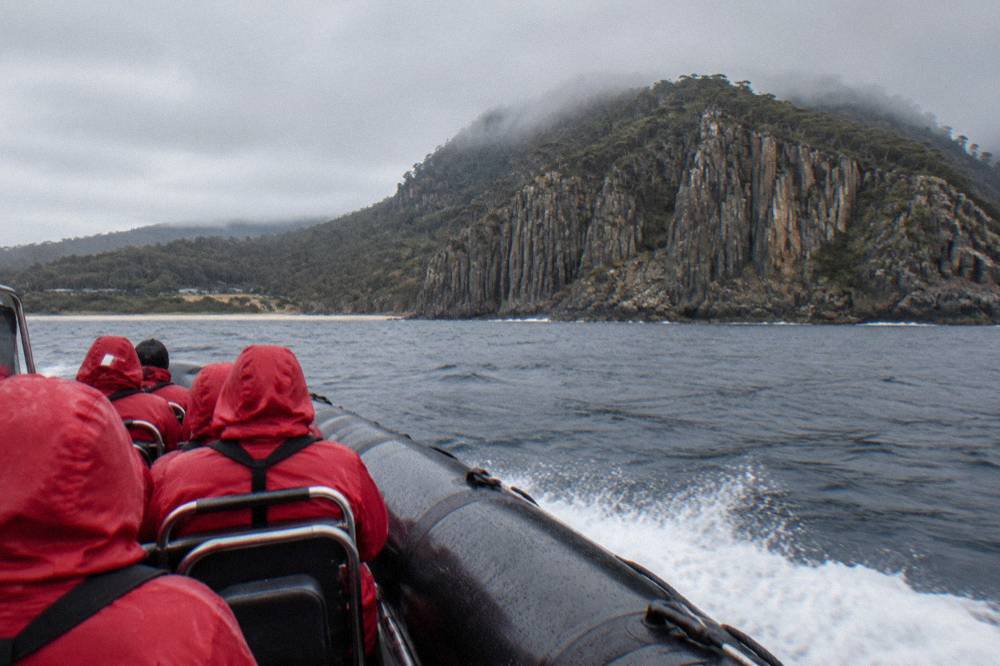 Passengers in red waterproof jackets seated in an open boat heading toward sea cliffs