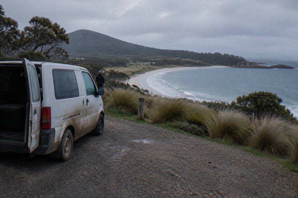 A small van parked at a coastal lookout above a curved white sand beach