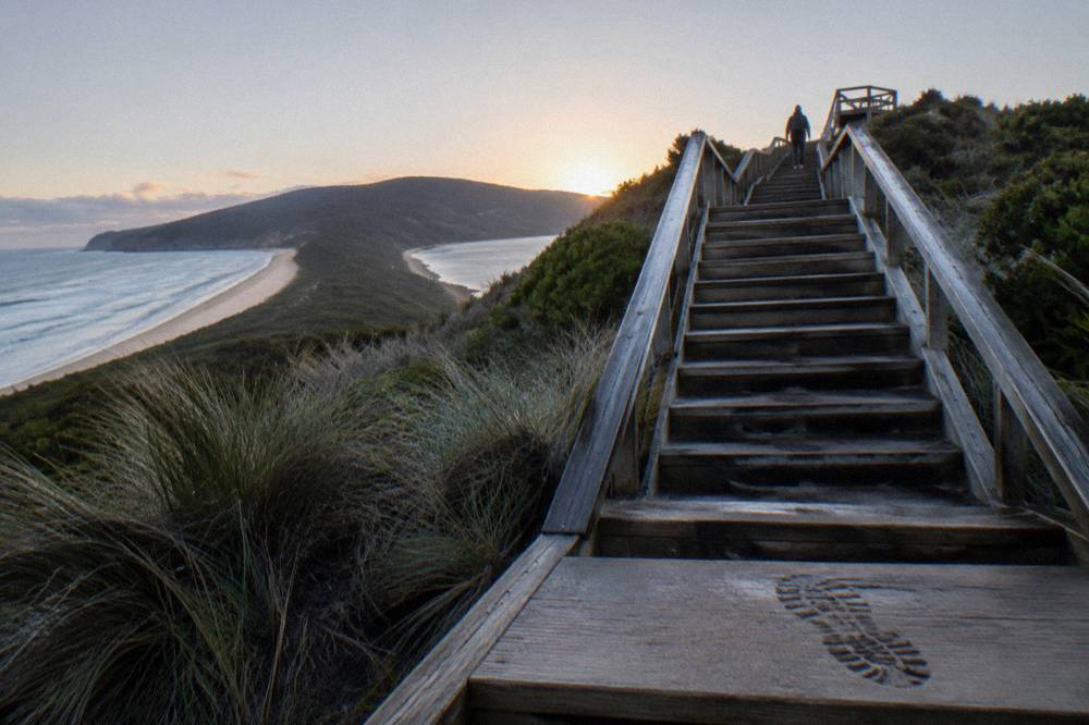 A wooden stairway climbing to a lookout platform above a narrow sand isthmus