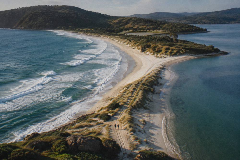 An aerial view of a narrow sandy isthmus connecting two green sections of an island with ocean on both sides