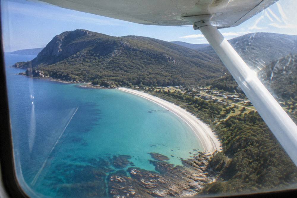A light aircraft wing above turquoise water and a long white beach along a forested coast