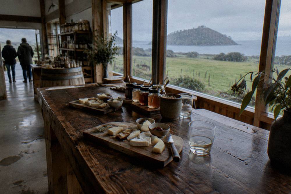 A wooden tasting counter with cheese boards, honey jars and glass tumblers arranged in front of a window overlooking green farmland