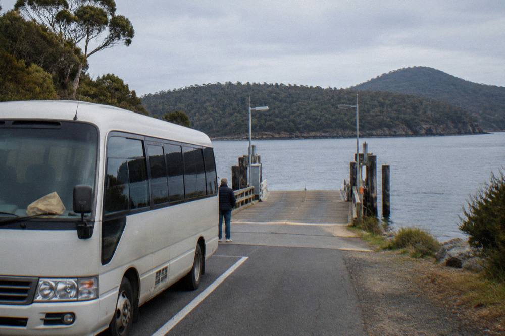 An air-conditioned minibus with tinted windows waiting at a small ferry terminal queue beside calm grey water
