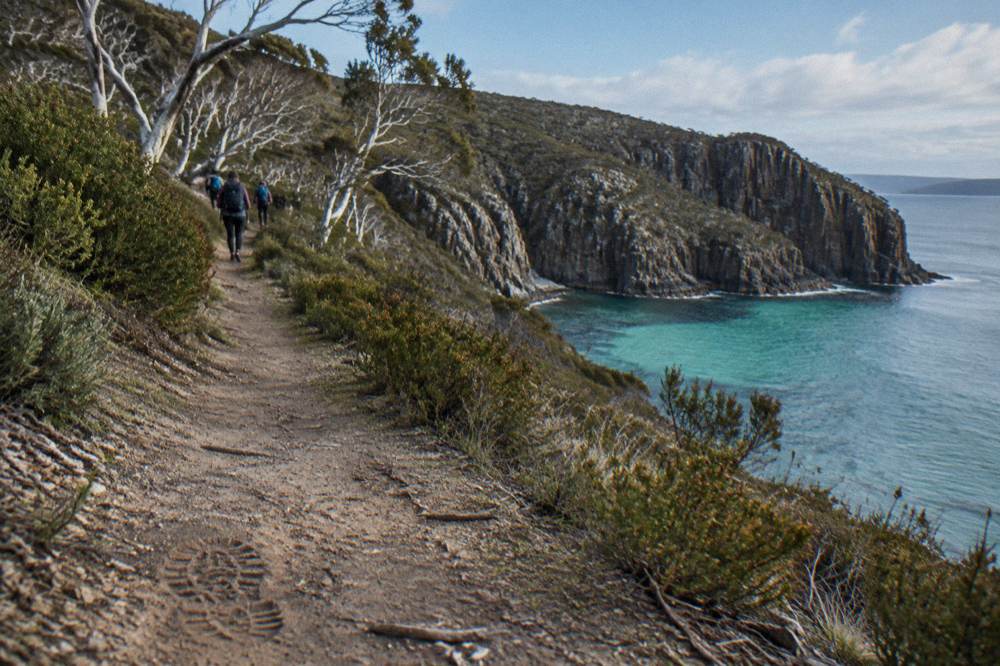 A small group of hikers with daypacks walking a coastal path above a turquoise bay and sea cliffs