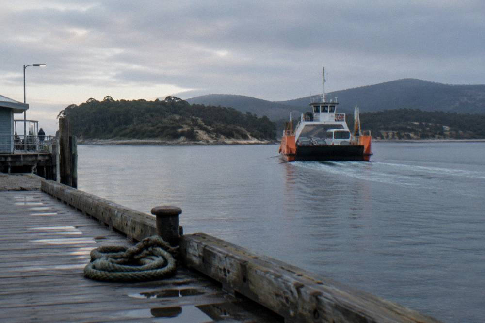 A vehicle ferry loaded with cars and a small bus easing away from a terminal jetty across calm water