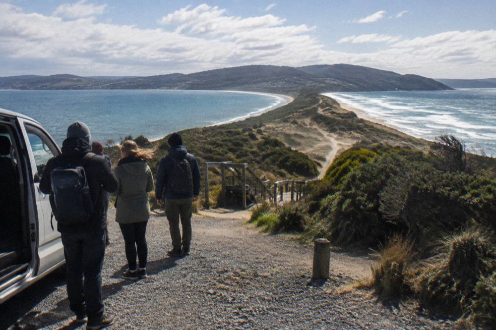 A guide standing beside an open van door speaking with a few travellers at a lookout above a long beach and headland