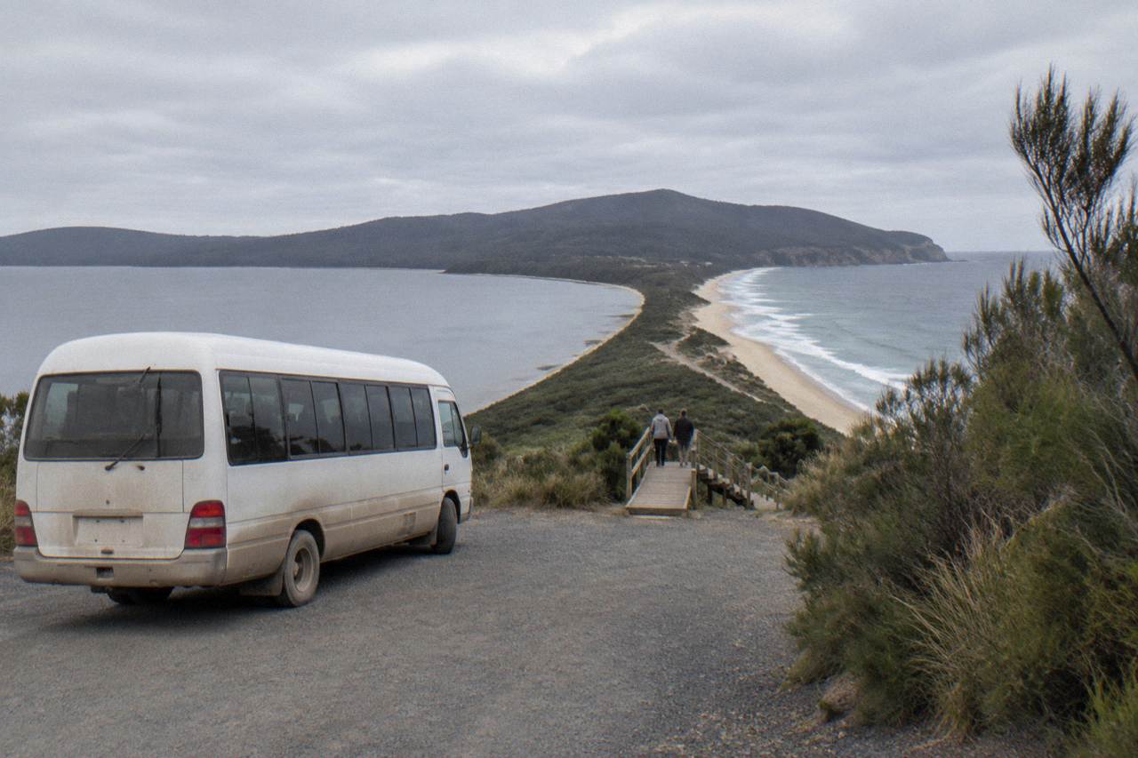 A small minibus parked beside a gravel turnout overlooking a narrow sandy isthmus joining two green island headlands under a cloudy sky