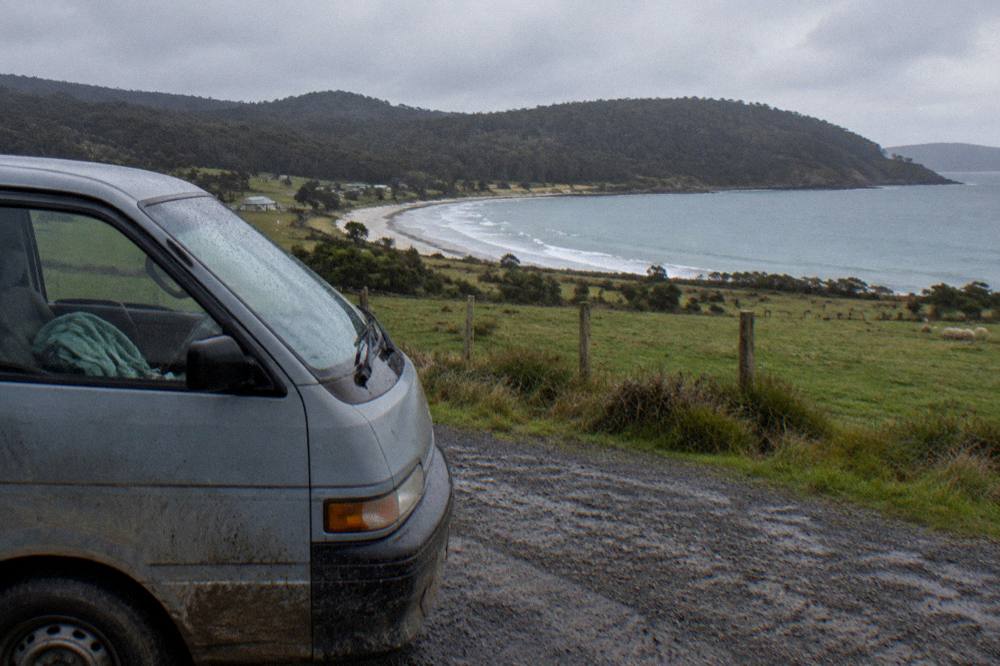 A small van parked on a gravel roadside overlooking a sheltered bay and farmland