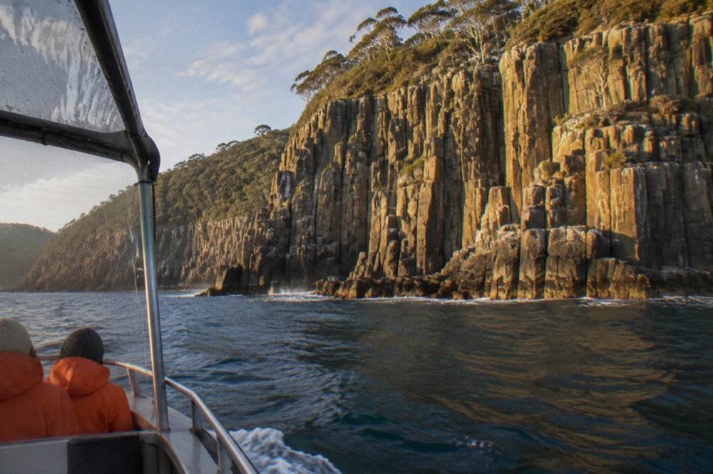 An open cruise boat travelling along tall sea cliffs and rock formations on a wilderness coastline