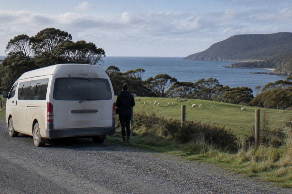 A minibus parked beside a coastal road with green farmland and ocean beyond