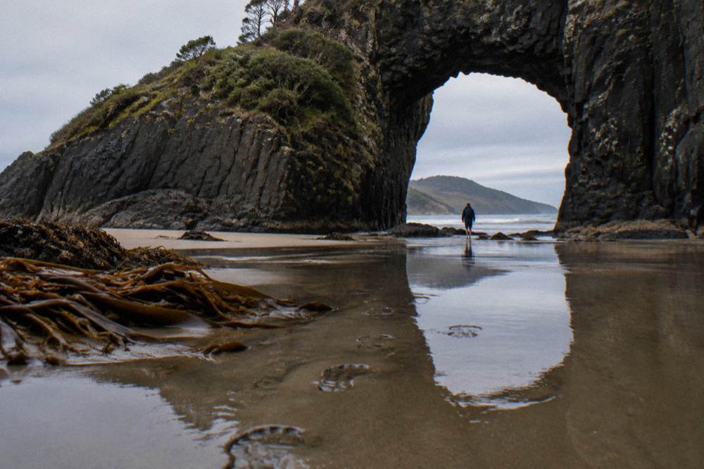 A natural rock arch in a cliff face at the edge of a beach with wet sand reflecting the sky