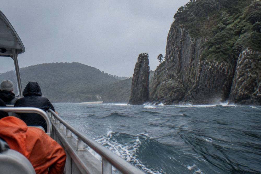 Open cruise boat cutting through swell beside tall sea cliffs with spray in the air