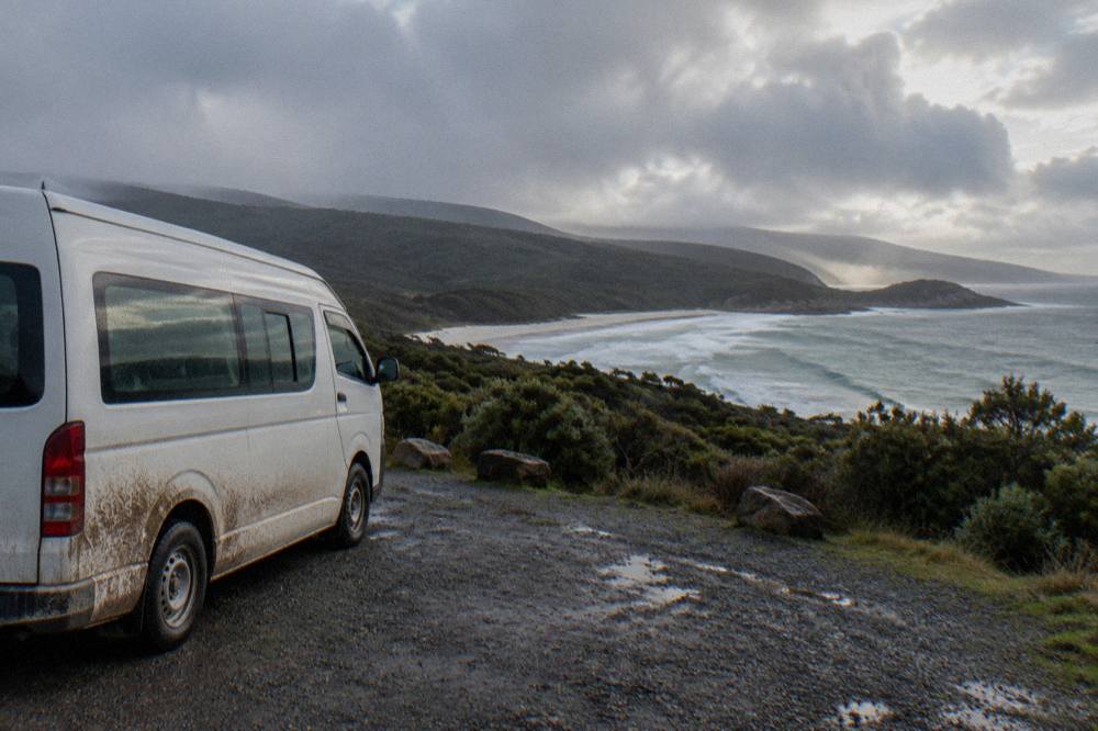 A minibus parked beside a coastal lookout with green hills meeting the sea