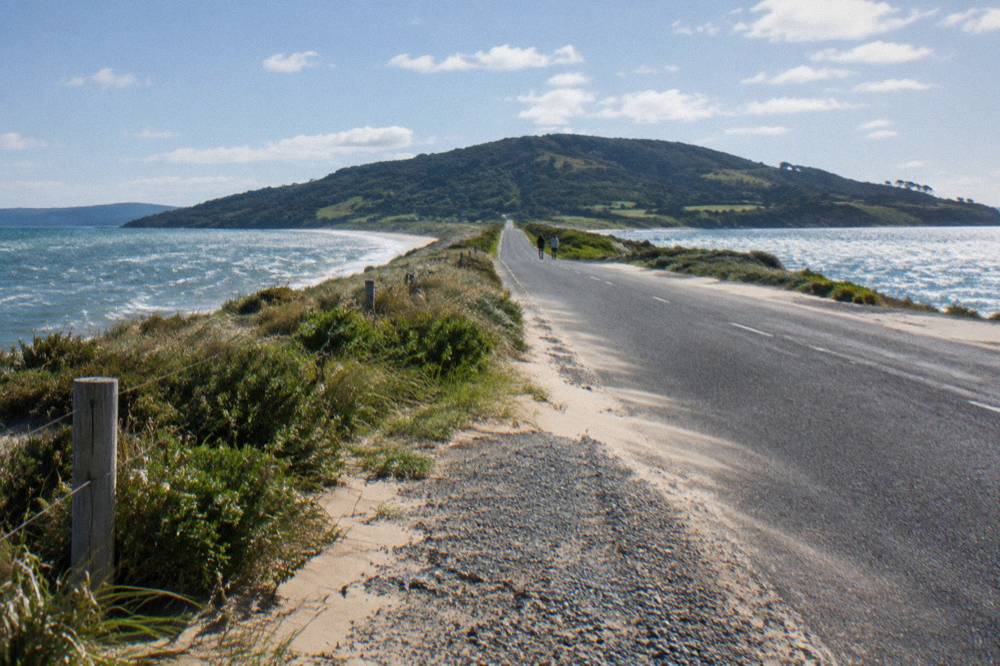 A quiet island road crossing low dunes toward a distant line of hills, water glinting on both sides