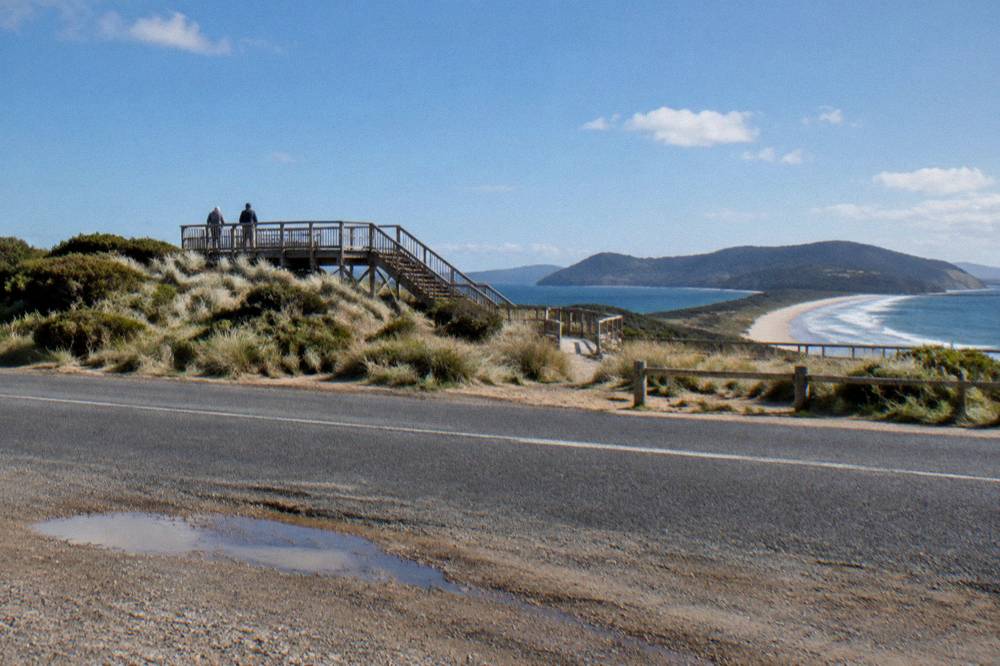 A viewing platform and staircase above dunes at midday, seen from a parking area beside an island road