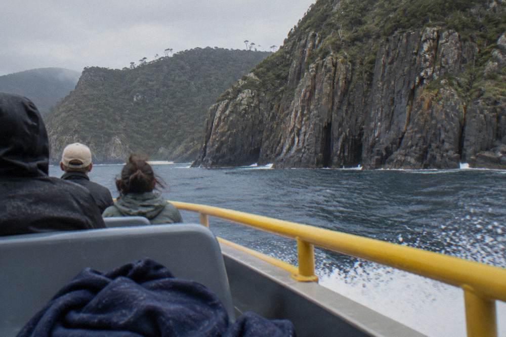 An open boat cruising close beneath tall columned sea cliffs above dark blue water.