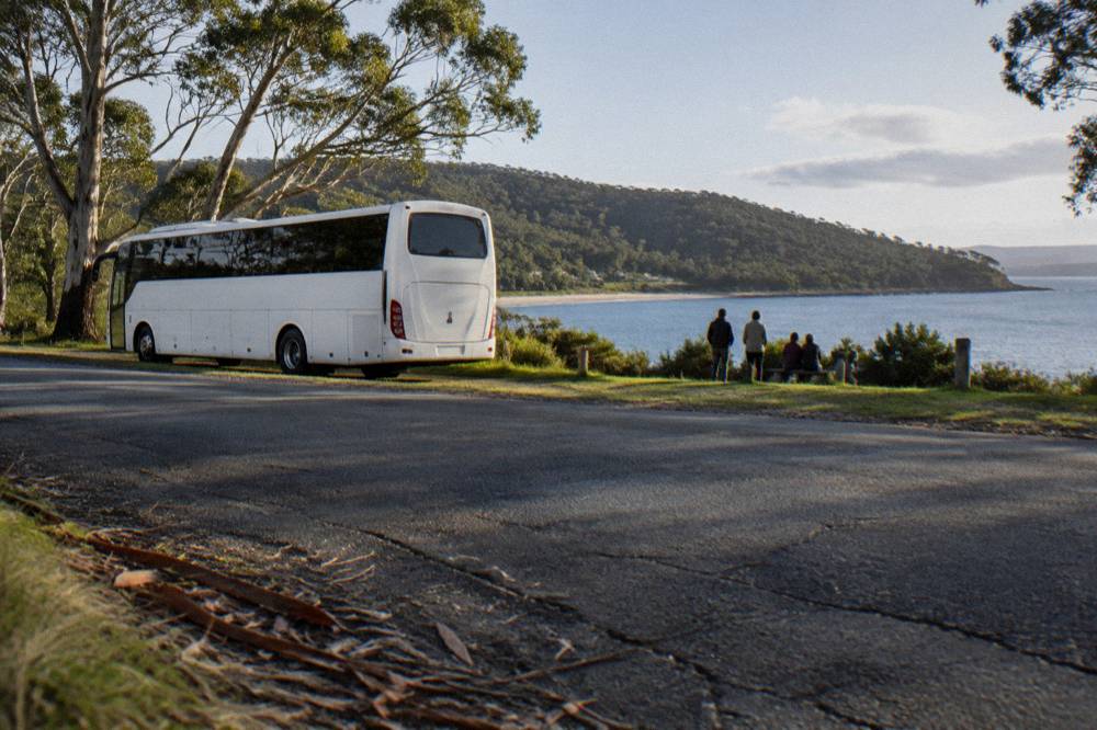 A coach parked beside a shorefront road with a calm bay and forested hills beyond.